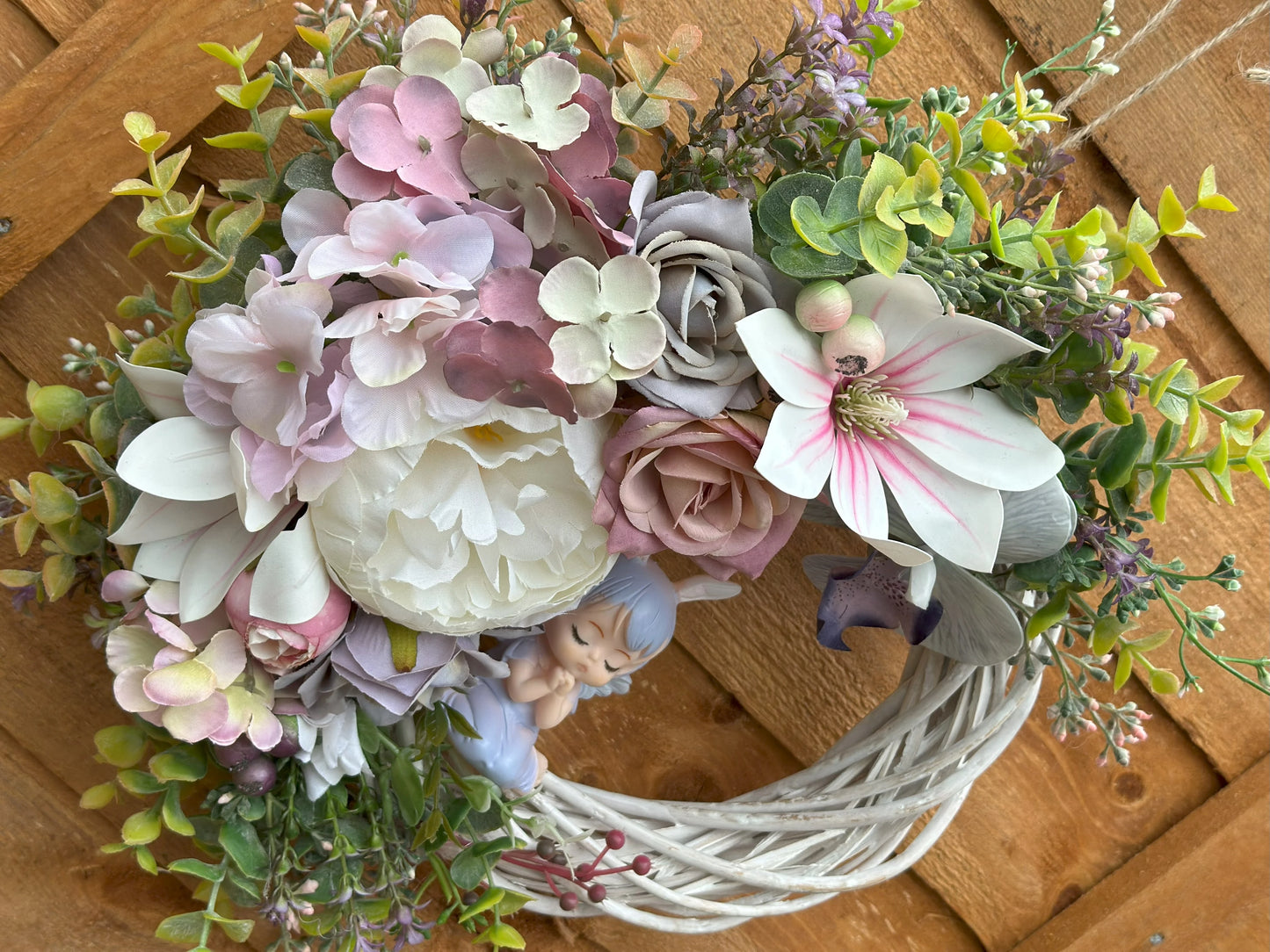 Handcrafted floral wreath with an angel figurine, featuring soft pink and white roses, green foliage, and a rustic white vine base.