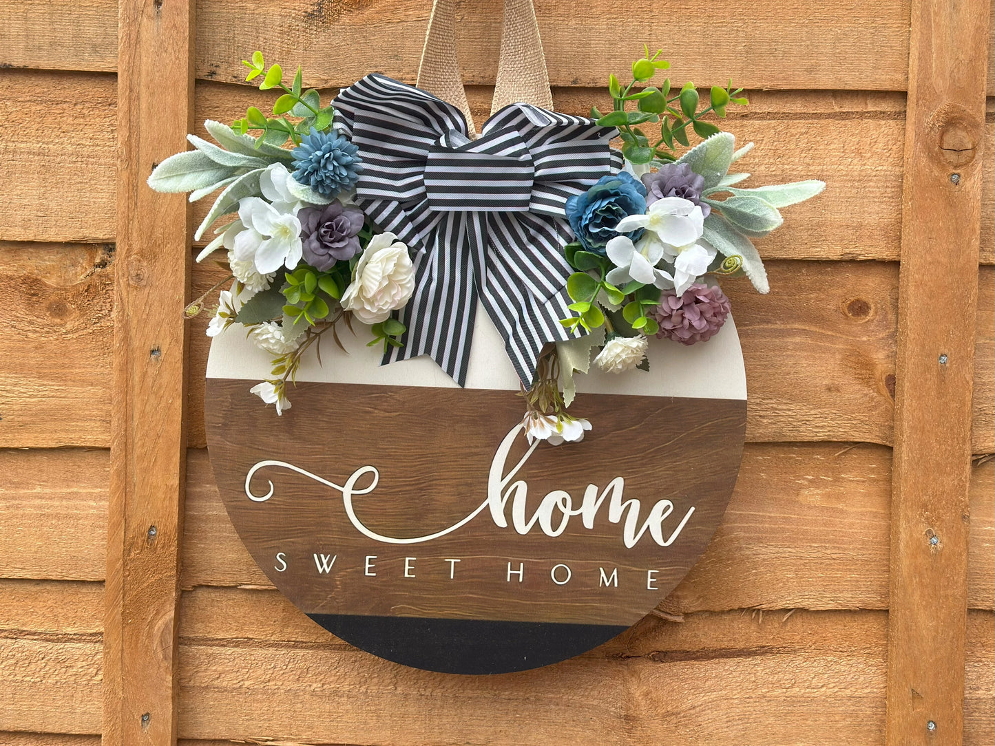 Handmade wooden wreath with artificial flowers and striped fabric bow, "Home Sweet Home" sign, rustic farmhouse door decor, modern classic wall hanging, cozy home decoration, indoor and outdoor wreath, neutral white and light brown tones.