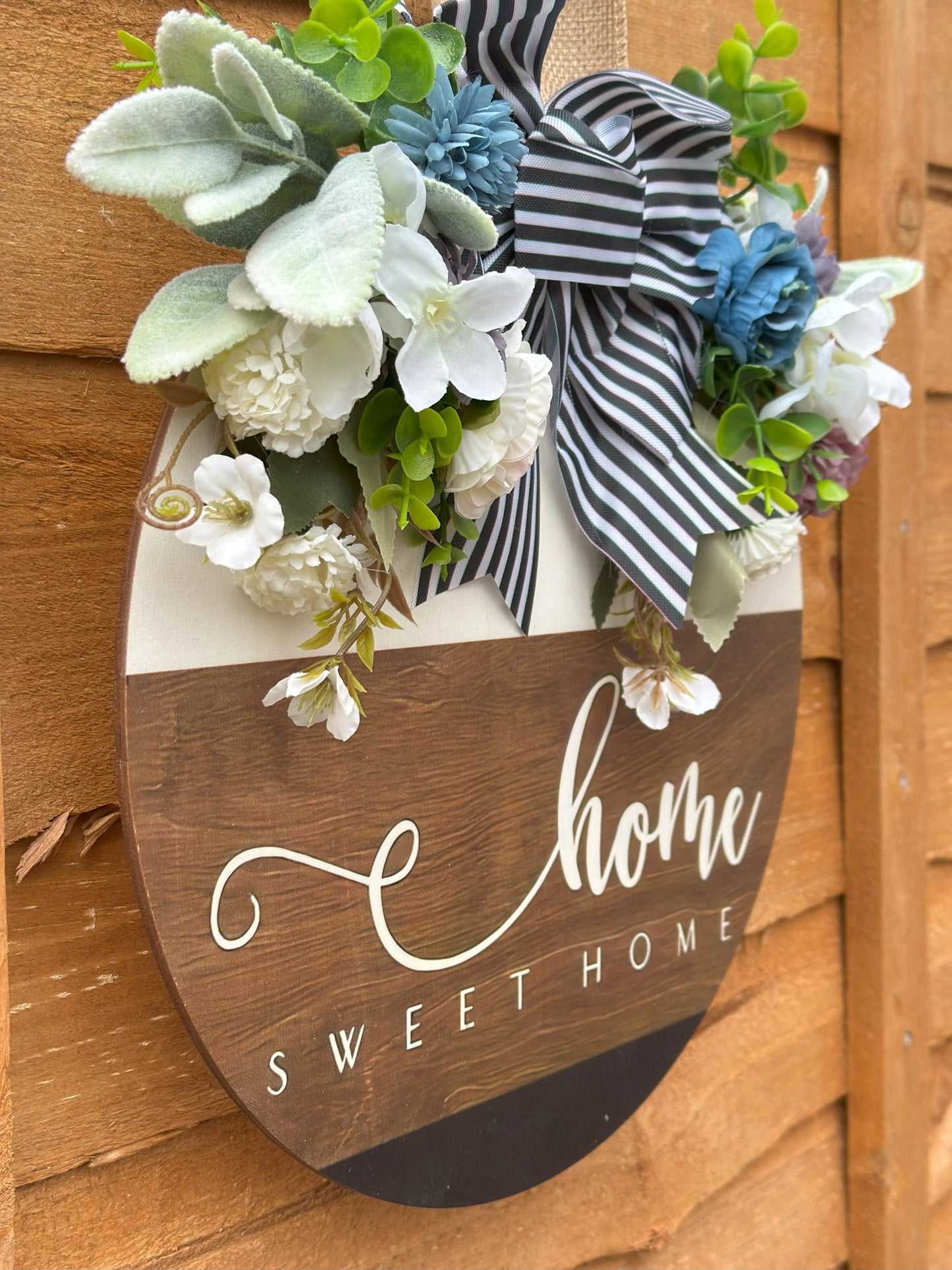 Handmade wooden wreath with artificial flowers and striped fabric bow, "Home Sweet Home" sign, rustic farmhouse door decor, modern classic wall hanging, cozy home decoration, indoor and outdoor wreath, neutral white and light brown tones.