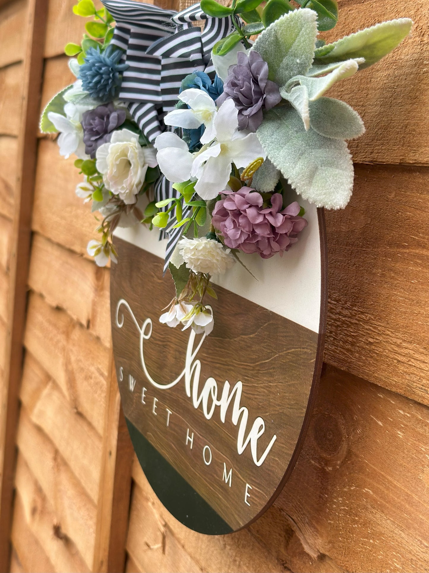 Handmade wooden wreath with artificial flowers and striped fabric bow, "Home Sweet Home" sign, rustic farmhouse door decor, modern classic wall hanging, cozy home decoration, indoor and outdoor wreath, neutral white and light brown tones.