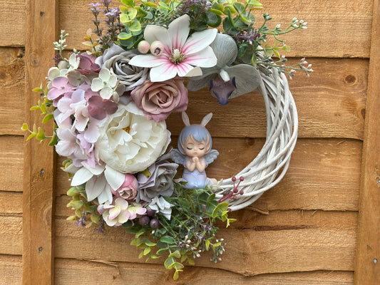 Handmade angel wreath with pastel roses and orchids on a white woven base, featuring a delicate praying angel figurine. Perfect for home decor.